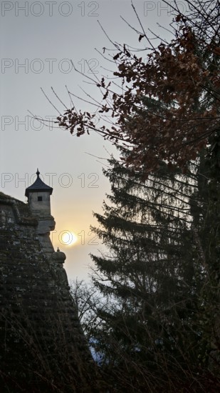 A castle tower, bay window of a fortress wall rising into the sky, surrounded by trees at sunset,