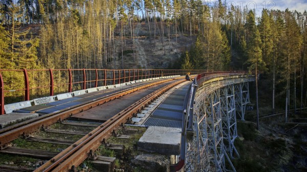 A person sits on the tracks of an old metal railroad bridge that leads through a forest,