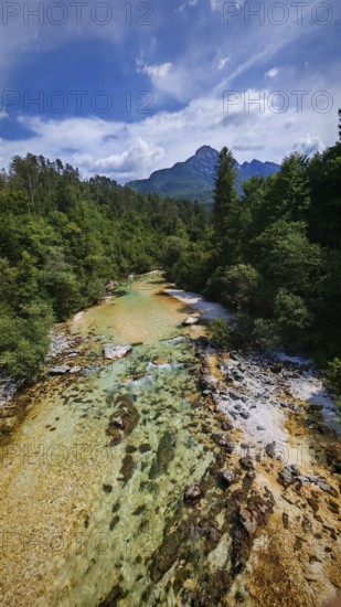A clear river flows through a forest with Triglav mountains in the background under a blue sky,