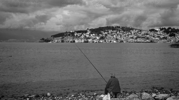 Black and white photo of a fisherman on a lake shore with a city in the background, Ohrid, North