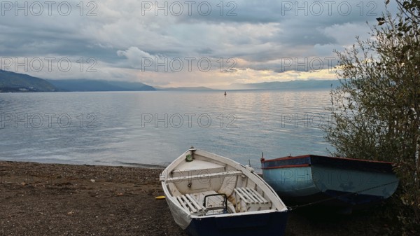 Two abandoned boats on the shore of a lake with mountains in the background under a cloudy sky,