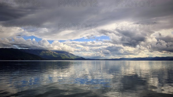Extensive lake with reflective, dramatic clouds and calm water and mountains in the background,