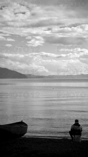 An angler is quietly looking at the shore of a lake, accompanied by a boat in black and white, Lake