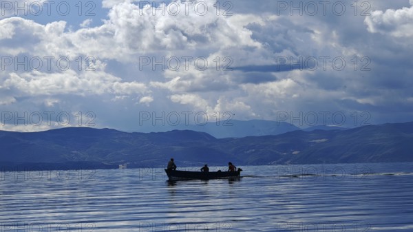 A small boat with silhouettes in a calm lake surrounded by mountains and dramatic cloud formations,
