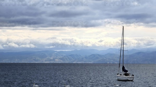 A sailboat on a calm sea with mountains in the background and cloudy sky, Lake Ohrid, North