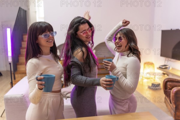 Three happy young women friends wearing sunglasses and casual clothes dancing, holding mugs, and