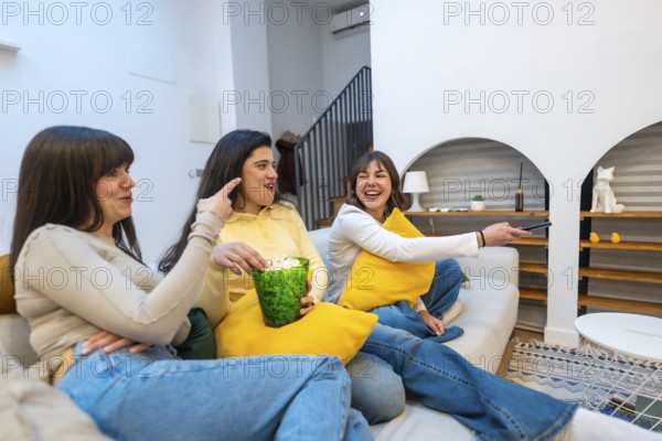 Three women on a sofa sharing popcorn, laughing and watching tv together in a cozy living room,