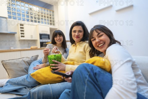 Three young women laughing and relaxed on a couch at home, enjoying a cozy movie night together