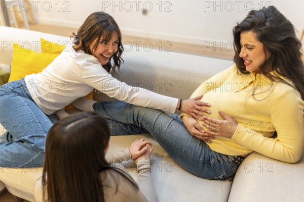 Two friends sharing a tender moment, sitting on a sofa and happily touching the belly of a smiling