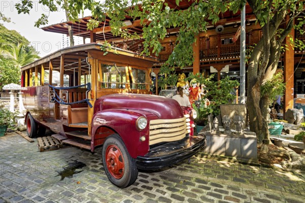 Red antique vehicle in front of a wooden building in tropical surroundings, surrounded by trees,