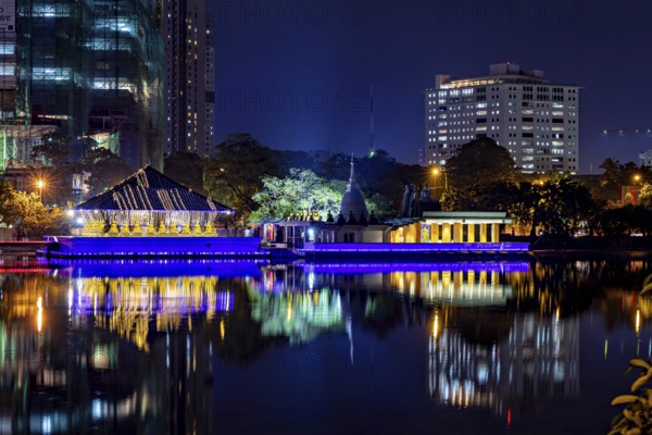 At night, a brightly lit temple is reflected in the calm waters of an urban lake, The Seema Malaka