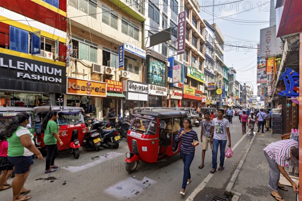 Bustling shopping street with rickshaws and pedestrians flanked by colorful buildings, the hustle