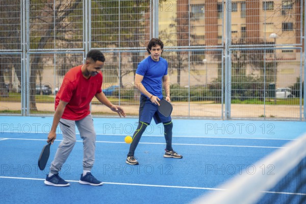 Two diverse young men compete on a blue outdoor pickleball court, focused and energetic as they