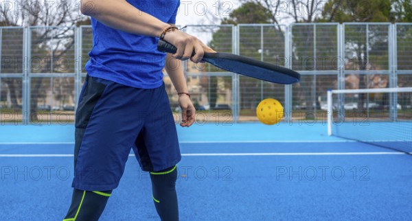 Athlete striking a yellow pickleball with a paddle on a vibrant blue outdoor court, mid rally