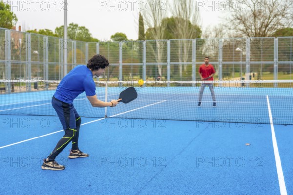 Two young men actively engaging in a game of pickleball, one hitting the yellow ball with a paddle,