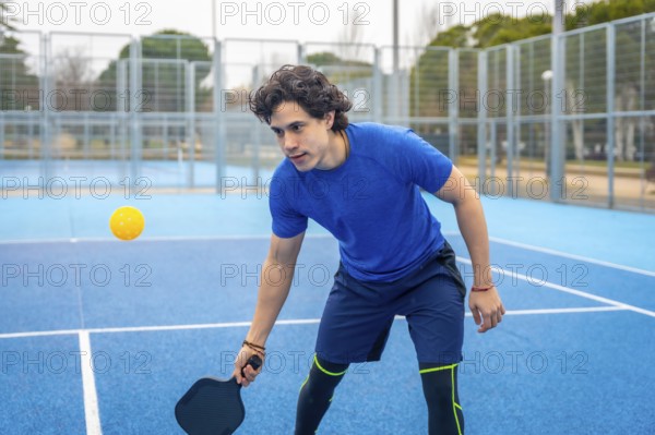 Young man holding a pickleball paddle, focused and ready to hit the yellow ball during an outdoor