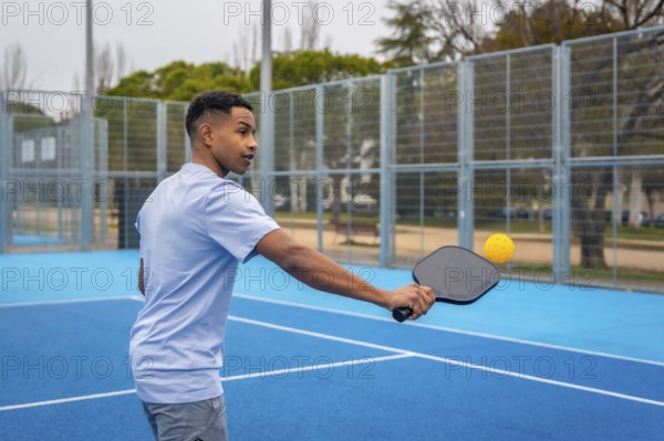 Young man wearing a light blue t shirt and grey shorts holding a paddle, hitting a yellow