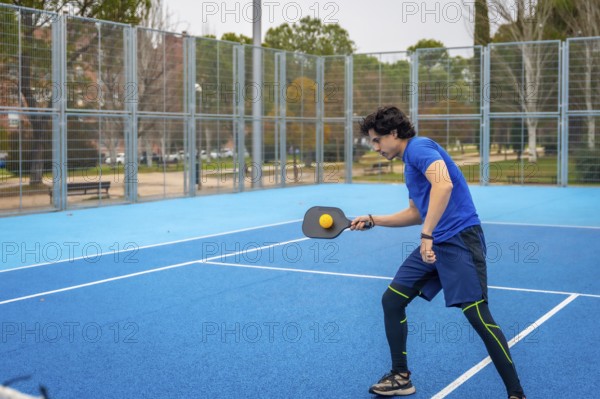 Young man in sportswear hitting a pickleball with a paddle on an outdoor blue court, focused and
