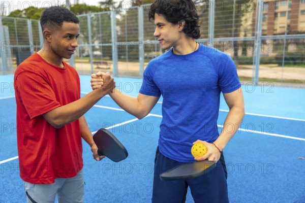 Two young diverse male athletes shaking hands on a blue pickleball court, holding paddles and a