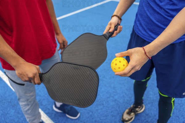 Two people are ready to play pickleball, holding paddles and a bright yellow wiffle ball on a