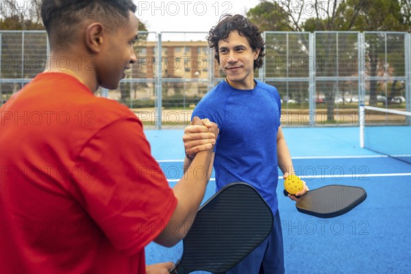 Two diverse male pickleball players shaking hands on a blue court, proudly displaying their paddles