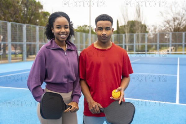 Young couple standing on a blue pickleball court, holding paddles and a ball, smiling at the