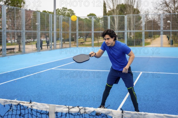 Young man in blue activewear playing pickleball on a blue court, intently hitting a yellow ball