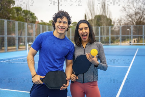Two young athletes smiling on a bright blue outdoor pickleball court, holding paddles and a ball,