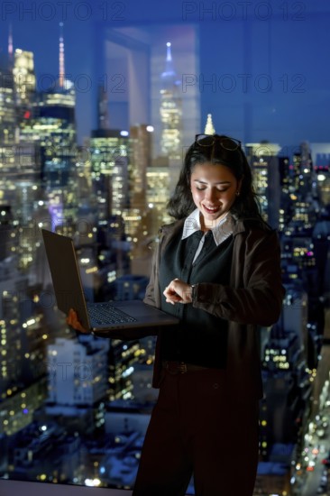 Young businesswoman standing by a large window, working on a laptop and checking her smartwatch