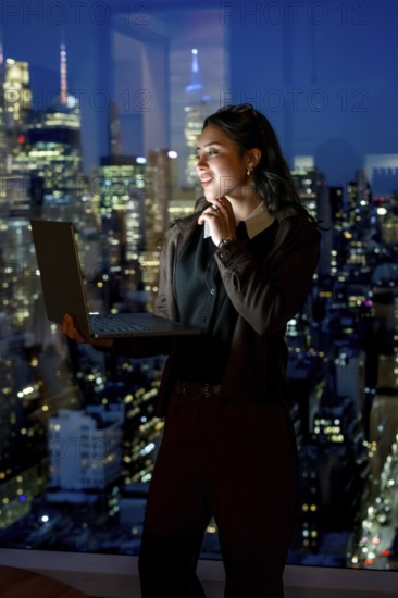 Young businesswoman standing confidently by a large window with her laptop, looking out at the