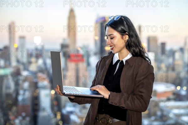 Young asian businesswoman using a laptop while standing against a blurred manhattan cityscape,