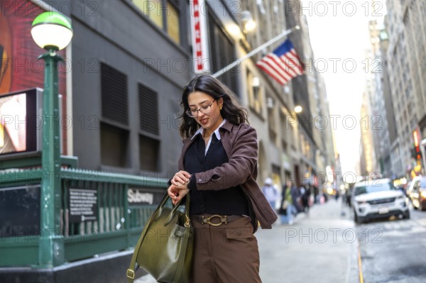 Young professional woman in a brown jacket checks her watch by a manhattan subway entrance, poised