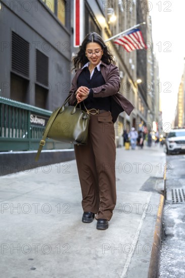 Young businesswoman walking urban street checking wrist watch while holding a handbag, representing