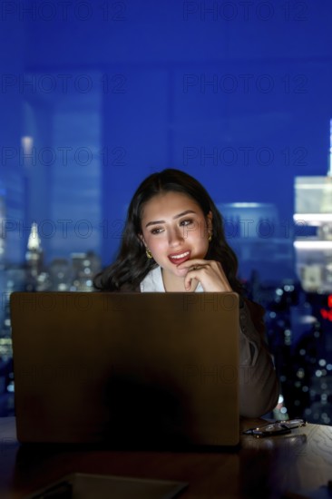 Young businesswoman working late on a laptop inside a modern manhattan office, illuminated by the