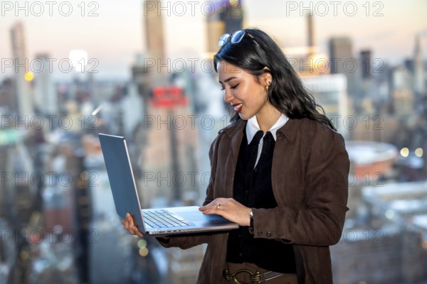 Young businesswoman stands on a manhattan rooftop, smiling and working on a laptop with the new