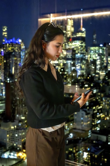 Young businesswoman standing by a window, engrossed in her smartphone, with the illuminated