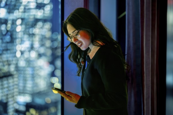 Young businesswoman standing by a skyscraper window at night, focusing on her mobile phone screen