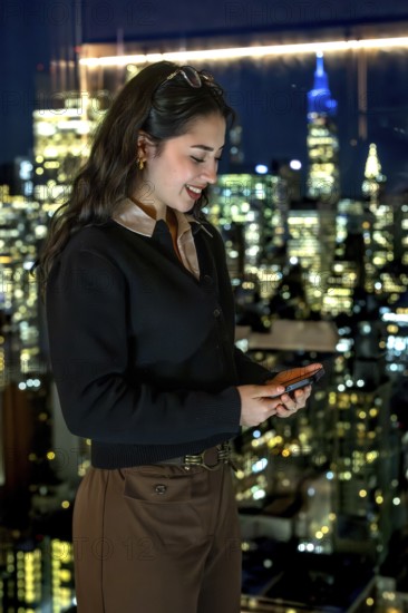 Young businesswoman smiling, looking at her smartphone while standing by a window with a stunning