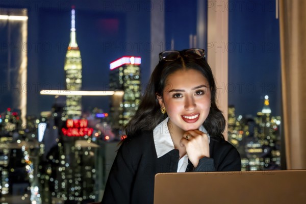 Young businesswoman smiling at camera, sitting in front of a window with a blurred night view of