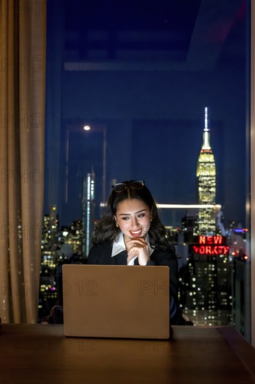 Young businesswoman observing laptop screen with a thoughtful smile, working late in a high rise