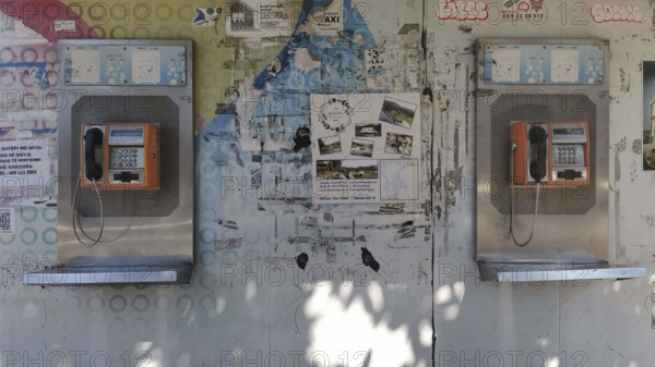 Two orange telephone boxes on a wall covered with graffiti and posters in an urban setting, Tirana,