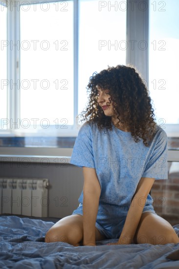 A young woman is sitting on a bed in a simple room. Bright daylight comes through the window,