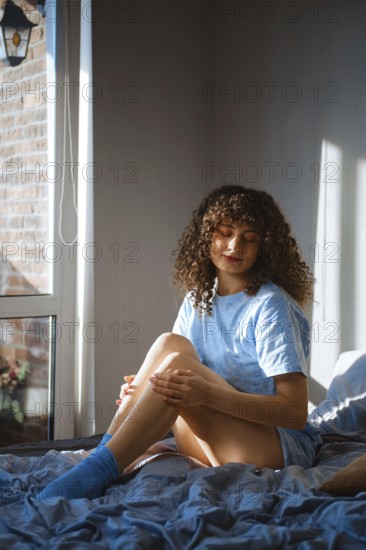 Dreamy woman is sitting on a bed near a window. She is looking down while touching her leg softly.
