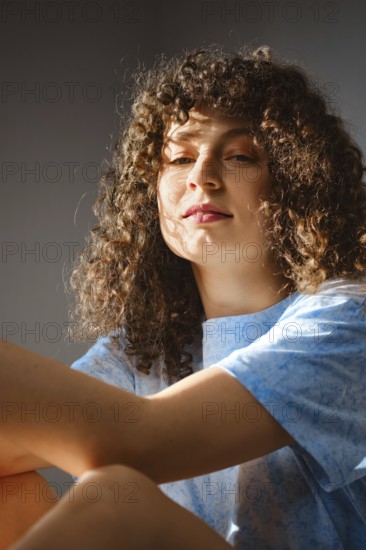 A young woman sits in a room, allowing sunlight to shine on her. She has curly hair and wears a