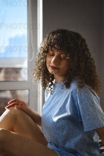 A woman sits near a window with natural light coming in. She has curly hair and is wearing a blue