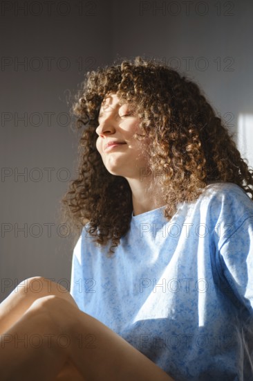 A woman sits relaxed by a window with sunlight on her face. She has curly hair and wears a blue