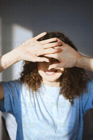 A woman stands in a well-lit room, smiling and covering her face with both hands to block sunlight.