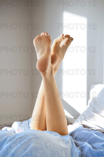 Crossed barefoot female legs on a bed. The legs raised in the air. Light comes in through a window,
