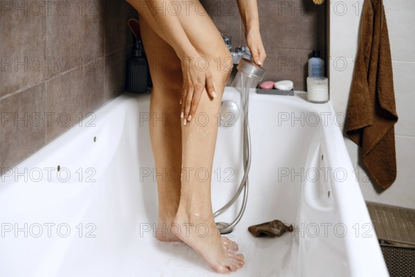 A woman stands in a bathtub washing her legs with a shower head. The bathroom has tiled walls,