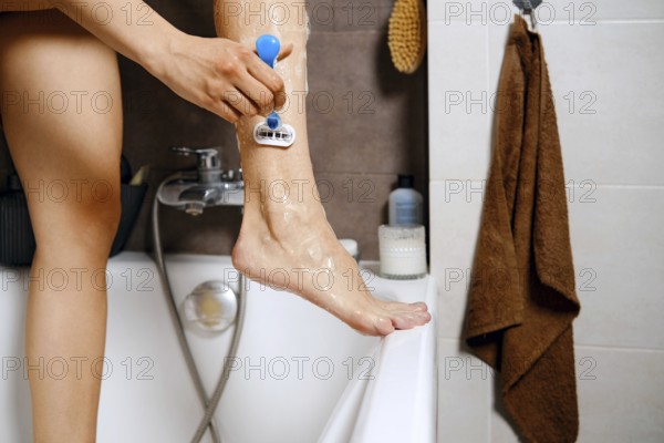 Unrecognizable woman is standing in a bathtub with one leg raised while using a razor to shave.